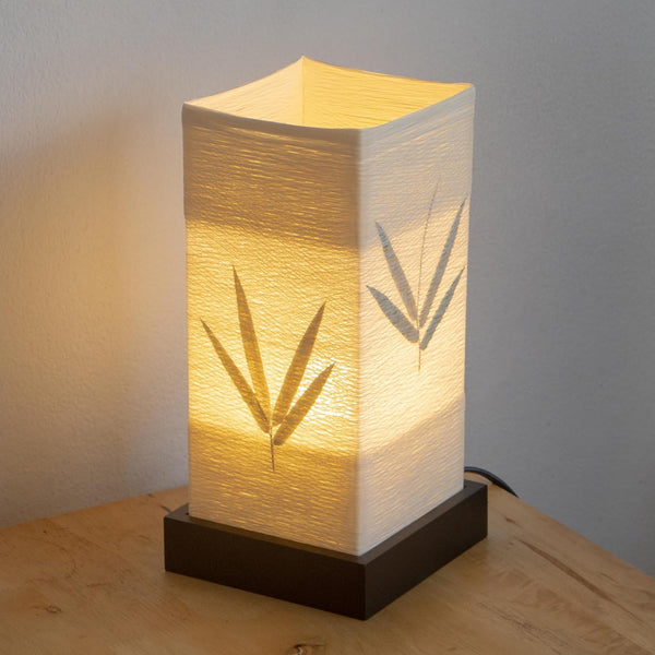 A paper lantern lit on a wooden base, bamboo leaves pressed into the shade