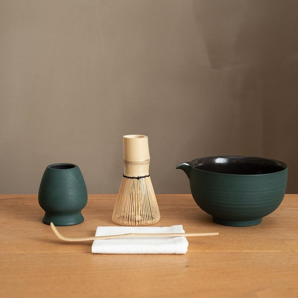 Matcha set on a wood table: a green chawan with a pouring spout, a bamboo whisk, a whisk holder, a bamboo scoop and a folded white tea cloth