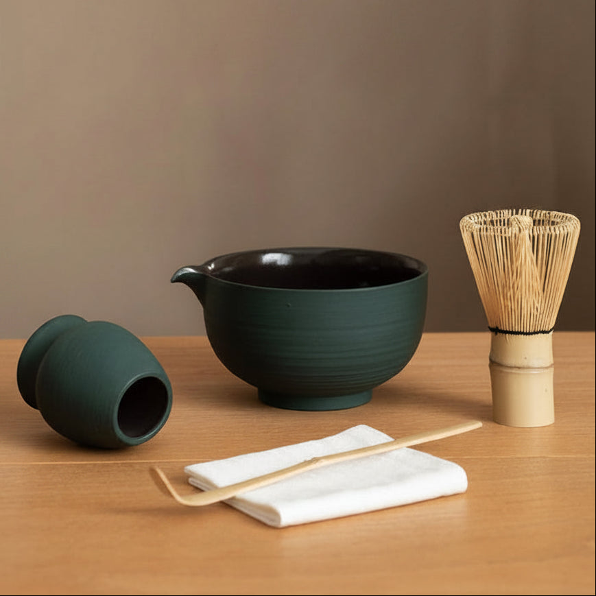 The set on a wood table: the green chawan with its pouring spout, the whisk holder tipped on its side, a bamboo whisk, a bamboo scoop and a folded tea cloth