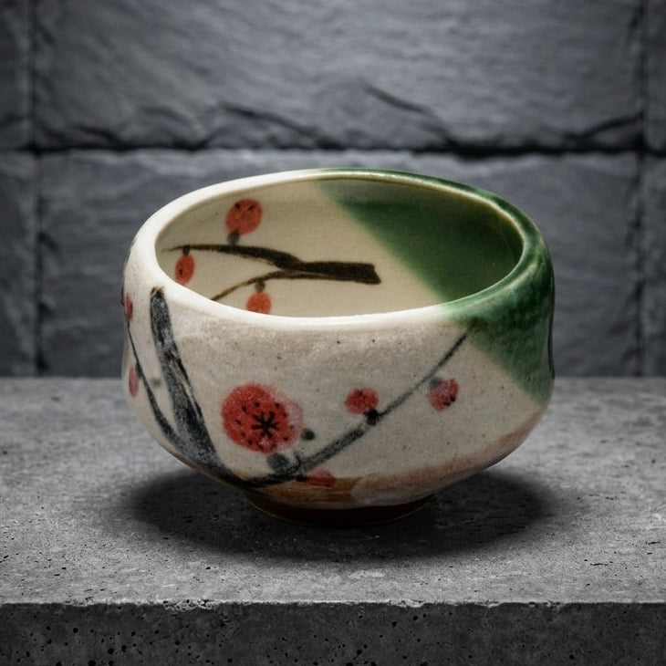 The chawan on a grey stone slab against dark slate: white glaze with red plum blossoms and a green glaze pouring over one side