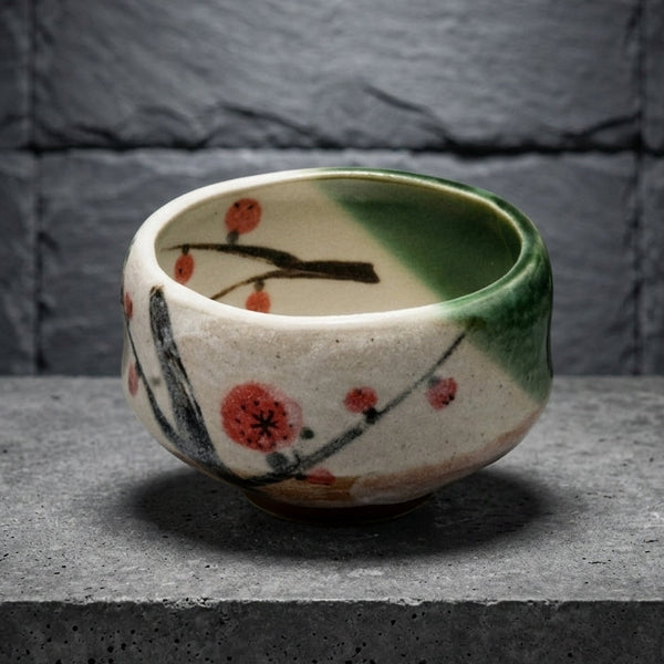 The chawan on a grey stone slab against dark slate: white glaze with red plum blossoms and a green glaze pouring over one side